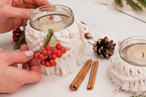 Mujer decorando un portavelas de vidrio en estilo macramé con ramas de abeto