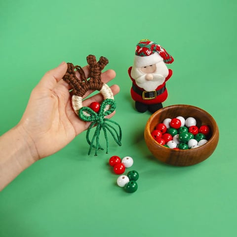 Mano sosteniendo adornos navideños en miniatura incluyendo un reno, una figura de Santa Claus y un cuenco de madera con cuentas rojas, blancas y verdes sobre fondo verde menta