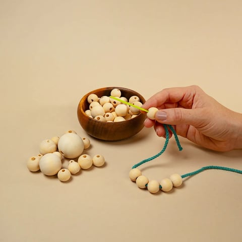 Mano sosteniendo cuentas de madera ensartadas en cordón turquesa junto a un bol de madera y cuentas dispersas sobre superficie beige