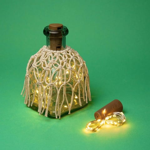 Botella de vidrio decorativa con cubierta de fibra natural tejida y luces de hilo blancas cálidas saliendo de ella sobre fondo verde