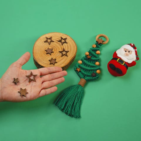 Mano sosteniendo estrellas de madera sobre un tablero redondo con patrones de estrellas, junto a un adorno navideño de árbol con cuentas y decoración de Santa sobre fondo verde