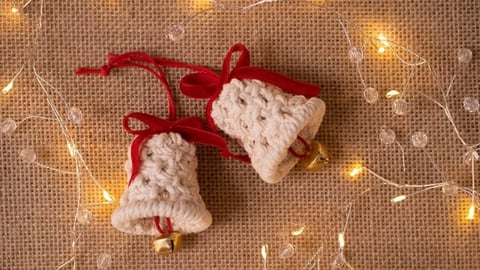 Dos campanas tejidas a crochet con cuerdas rojas sobre un fondo de tela tejida color canela con luces doradas de hadas
