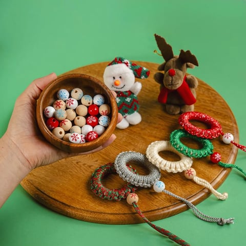 Mano sosteniendo un cuenco de madera lleno de cuentas de colores junto a un muñeco de nieve de fieltro y un reno sobre una bandeja de madera con pulseras de cuentas sobre fondo verde