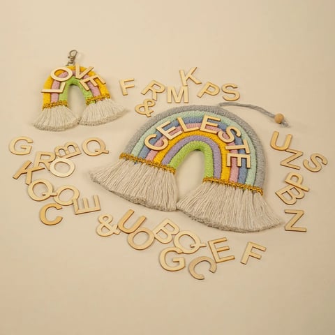 Dos adornos decorativos de tela en forma de arcoíris con flecos de macramé y fichas de letras de madera dispersas sobre fondo beige