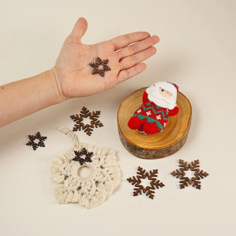 Mano sosteniendo adornos de copos de nieve color marrón con decoraciones navideñas y una figura pequeña de Santa Claus sobre superficies de madera
